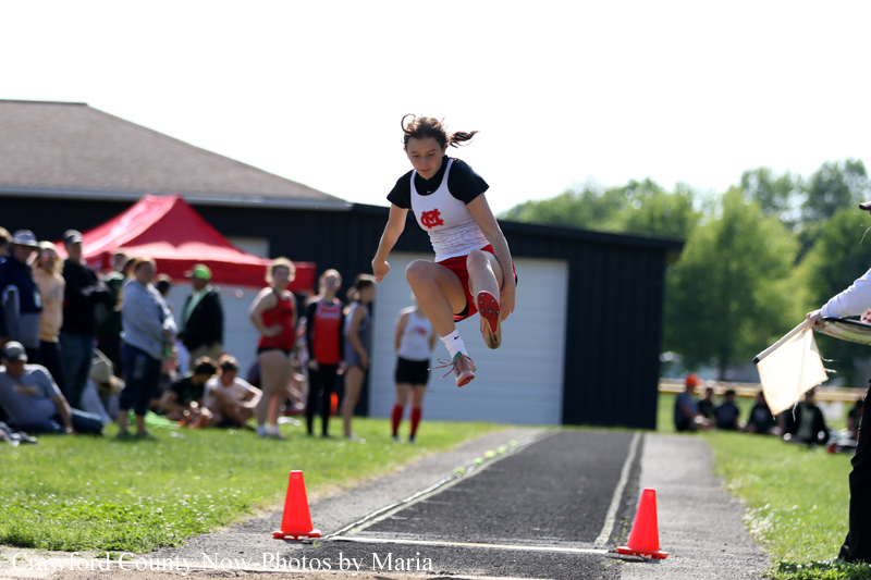 Young female track athlete in mid-air during a jump on a running track, with spectators watching in the background.