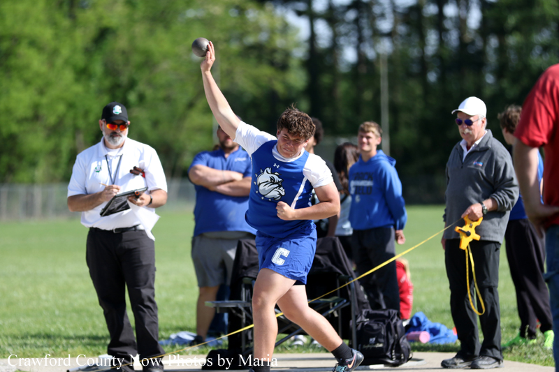 Male athlete in blue and white uniform launches a shot put while spectators and officials watch on a sunny field.