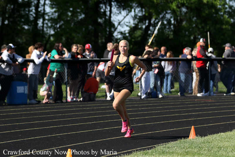 Female sprinter in black and yellow with pink shoes, holding a baton during a relay on a track while spectators watch from behind a fence.
