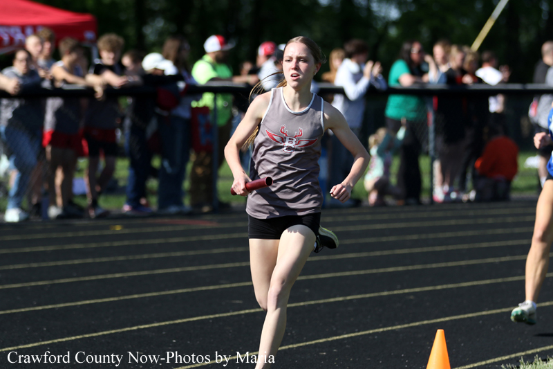 Female baton runner mid-stride on a track, holding a baton, with spectators behind a fence in the background.