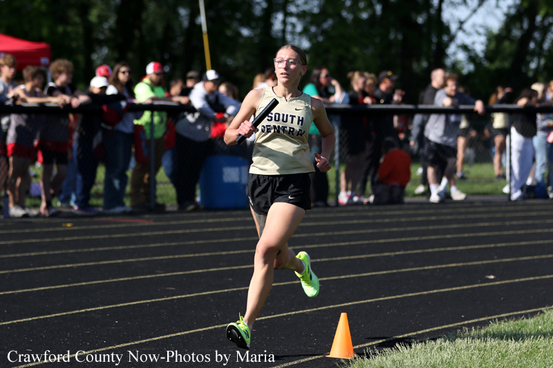 Female athlete sprinting on a track with a baton during a relay race, crowd watching behind a fence.