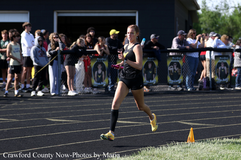 Female sprinter in black uniform running on a track with yellow shoes, spectators behind a fence watching the race.