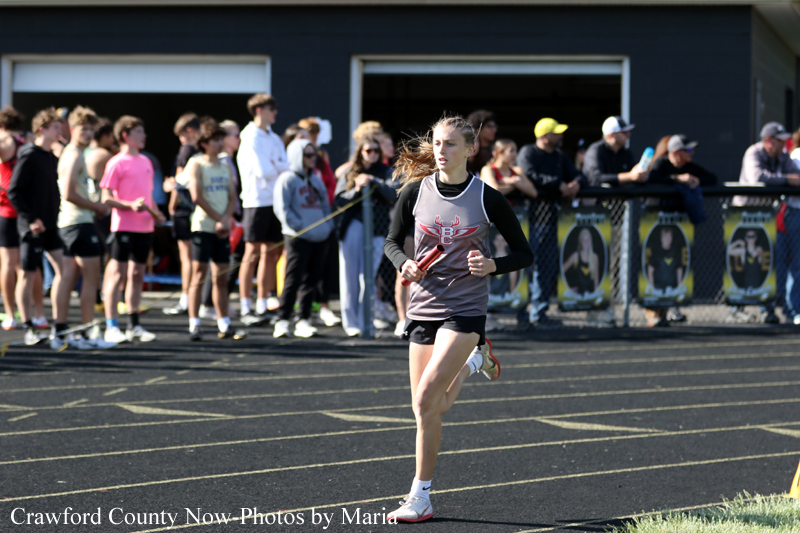 Girl in a gray relay uniform sprinting on a track, baton in hand, with a cheering crowd in the background.