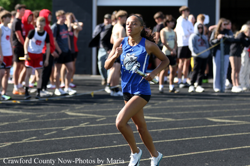 Female runner in a blue relay uniform holding a baton, sprinting on a track with a crowd of spectators in the background.