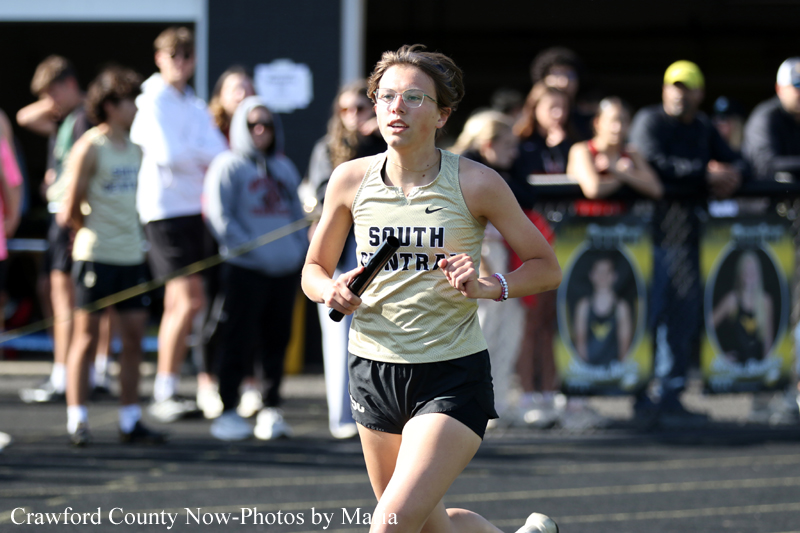 Female runner in a beige tank top and black shorts sprinting with a baton during a relay, spectators in the background.