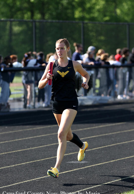 Female track sprinter running on a track with a pink baton, wearing a black kit with a gold eagle emblem as spectators watch behind a fence.