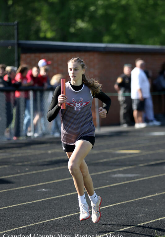Female student relay runner in a gray and black uniform holding a red baton on a track, with spectators behind the fence.
