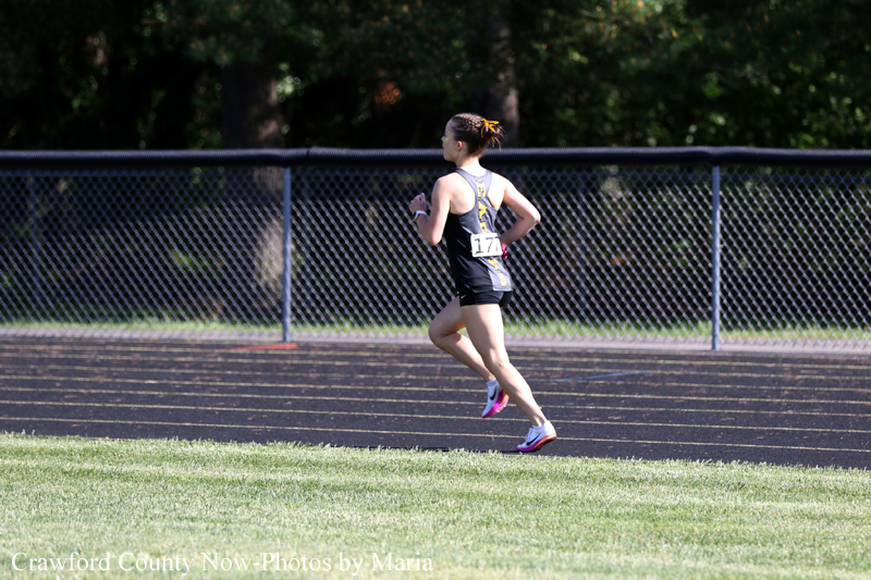 Female runner in black athletic outfit sprinting on an outdoor track with a chain-link fence in the background.