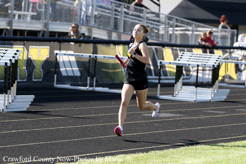 Female relay runner in a black uniform sprints on a track, holding a pink baton during a race.