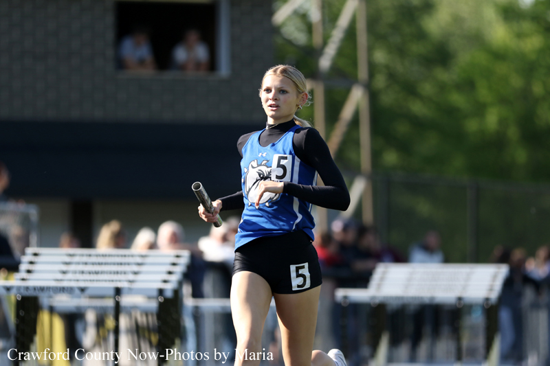 Female relay runner in a blue jersey with number 5, sprinting on an outdoor track with a baton in hand.