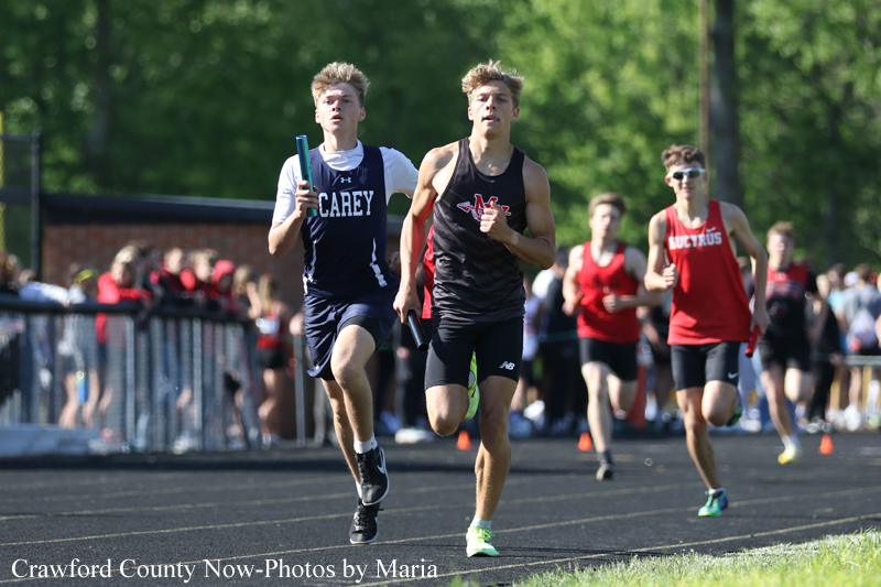 Two male sprinters lead a relay race on an outdoor track, one in a blue Carey uniform and the other in a black singlet, baton in hand.