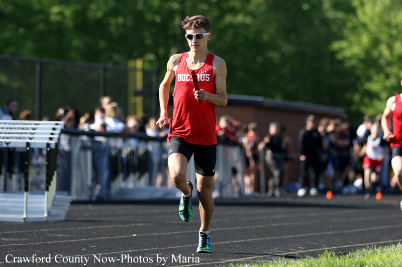 Male sprinter in a red sleeveless top and sunglasses runs on a track during a crowded meet.