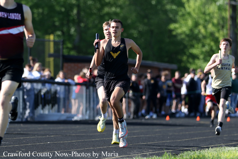 A male relay runner in a black uniform sprints on a track, holding a baton as runners race behind him with spectators in the background.
