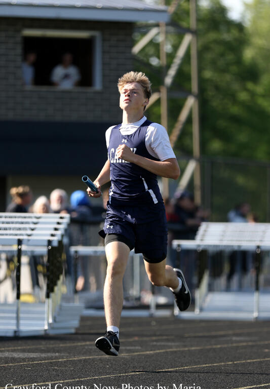 Male relay runner in navy and white sprinting on track with baton, spectators behind on a sunny day.