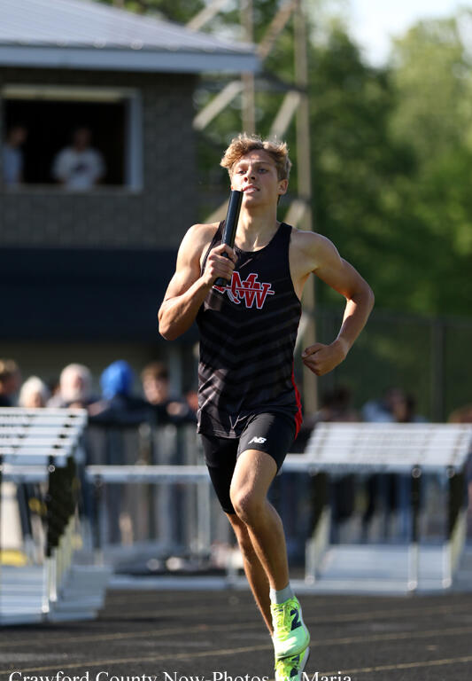 Male sprinter in a black and red uniform runs on a track holding a baton, with spectators and a scoreboard in the background.