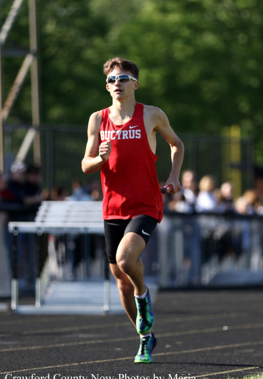 Male track runner in a red Bucyrus singlet sprinting on a track with spectators in the background, wearing sunglasses.