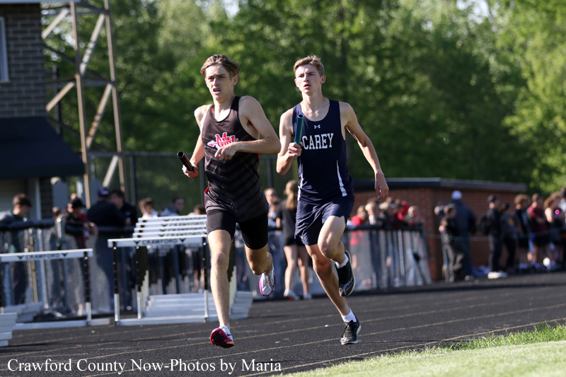 Two male sprinters race on an outdoor track, one in a black uniform and the other in navy with 'CAREY' on it, as spectators watch from the sidelines.