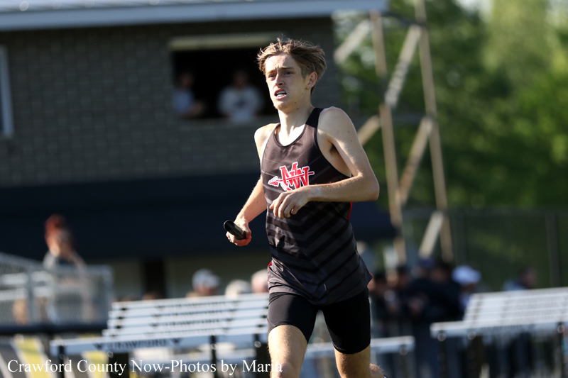 Male runner in a black singlet holding a baton during a relay on an outdoor track, with spectators in the background.