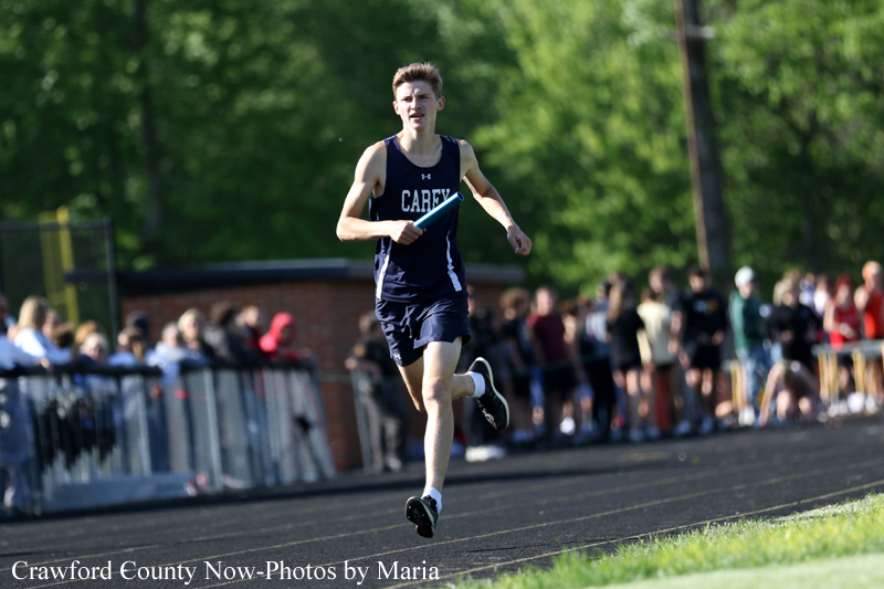 Male sprinter in navy uniform running on a track with a baton as spectators watch.