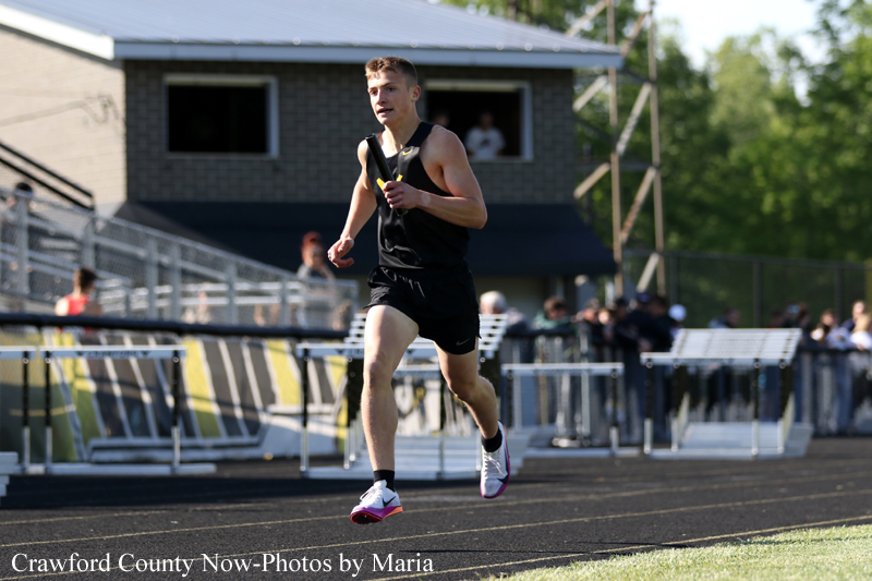Male sprinter in a black tank top and shorts runs on a track holding a baton during a relay race.