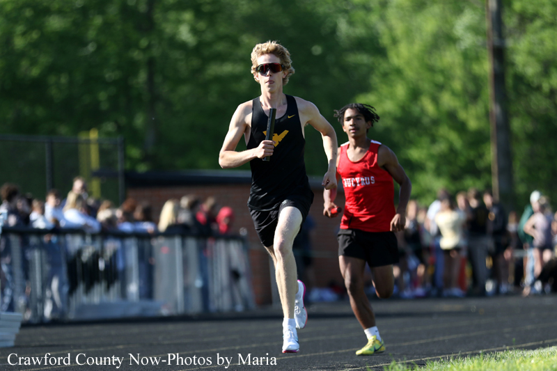 Two male sprinters race on an outdoor track; the lead runner wears a black singlet with sunglasses, chased by a competitor in red.