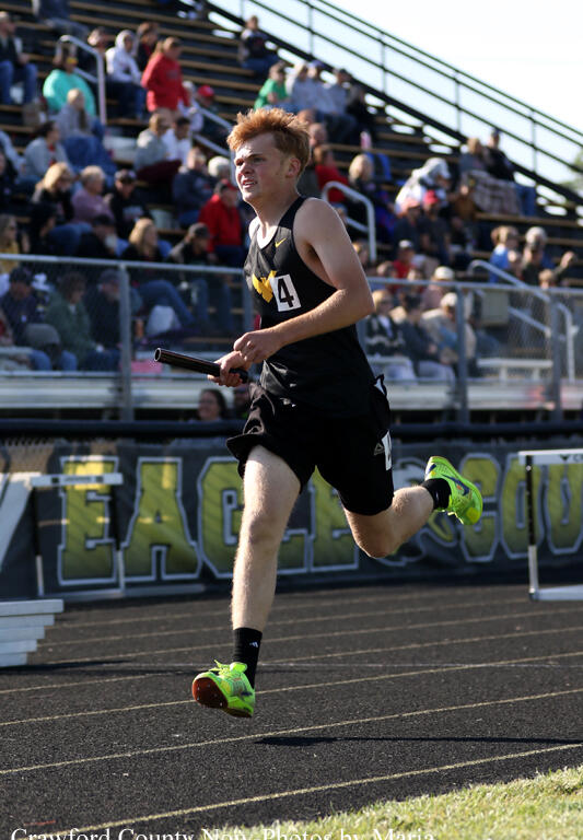 Relay race runner sprinting on a track with baton in hand; wearing black uniform with bib number 4 and neon green shoes, stands full of spectators in the background.