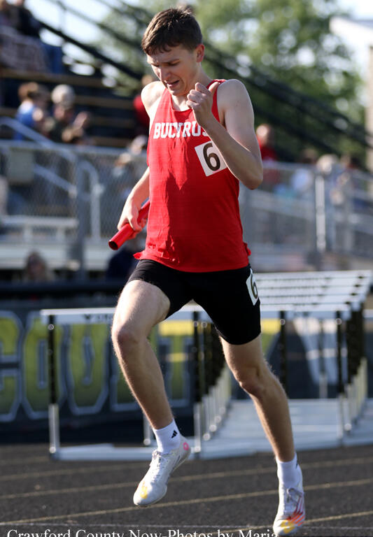 Male track athlete in a red singlet and black shorts jumps over a hurdle, holding a red relay baton in his left hand (bib number 6).