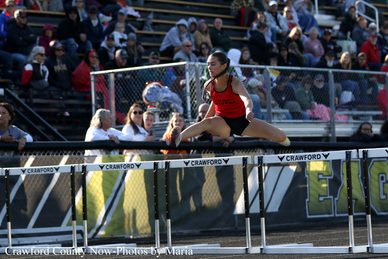 Female hurdler in red top clears a hurdle during a track race, stands against a crowd in the stands behind a chain-link fence.