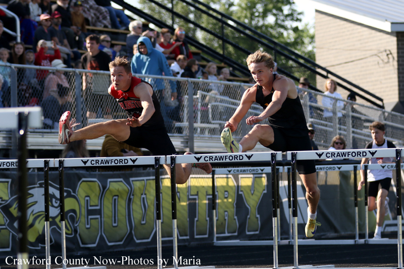 Two male track athletes in black uniforms leap over hurdles during a sprint on an outdoor track, with a crowd watching from the stands in the background.