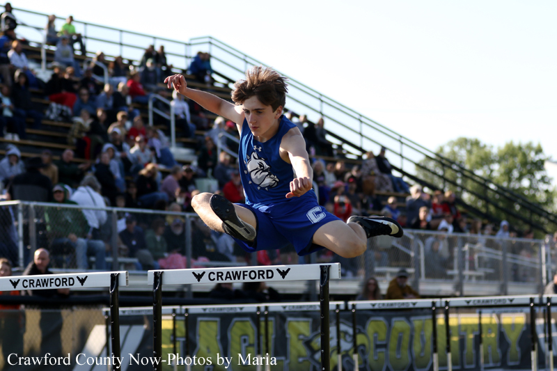 Male track athlete in a blue uniform mid-jump clearing a hurdle, with spectators in the stands behind him on a sunny day.