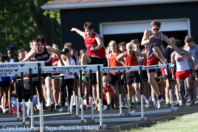 Several teenage runners leap over hurdles in a track race as a crowded group of spectators watches from behind the barriers.