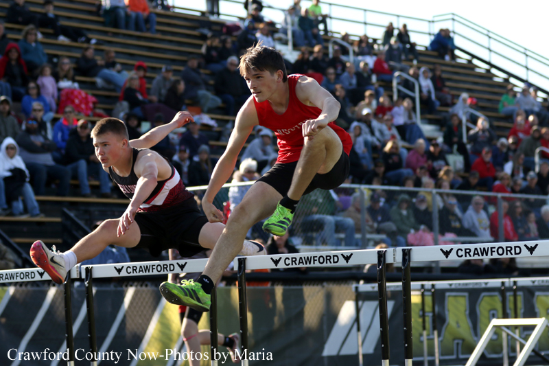 Two male hurdlers mid-air during a track race, leaping over hurdles as a crowded stadium watches from the stands.