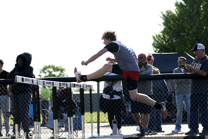 Male track athlete in gray jersey and red shorts clears a low barrier on a chain-link fence during a sunny track event, with spectators in the background.