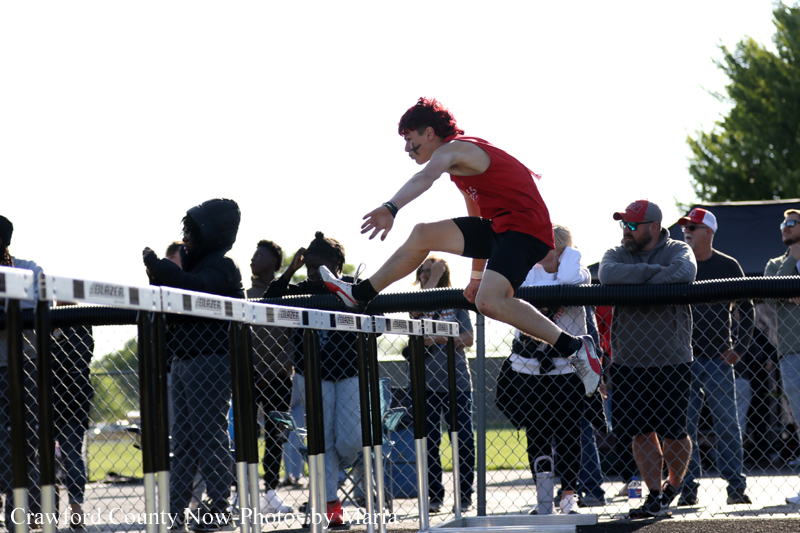 Male athlete in a red shirt leaps over a barrier on a track, with spectators watching behind the fence.