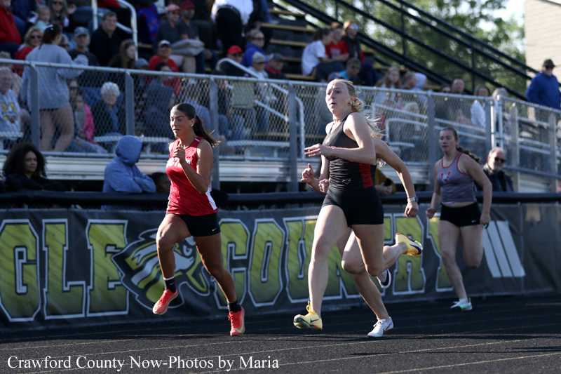 Three female runners sprint on a track as spectators watch from the stands below a chain-link fence.