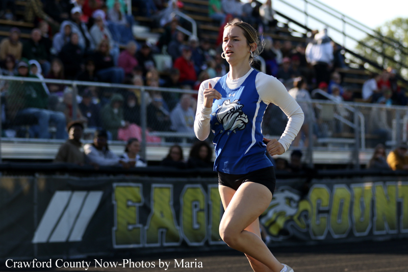 Female sprinter in a blue singlet with a bulldog logo, running on a track with stands and spectators behind her in a race.
