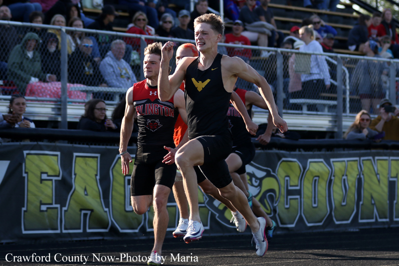 Two male sprinters race toward the finish line at a high school track meet, with a cheering crowd in the bleachers behind them.