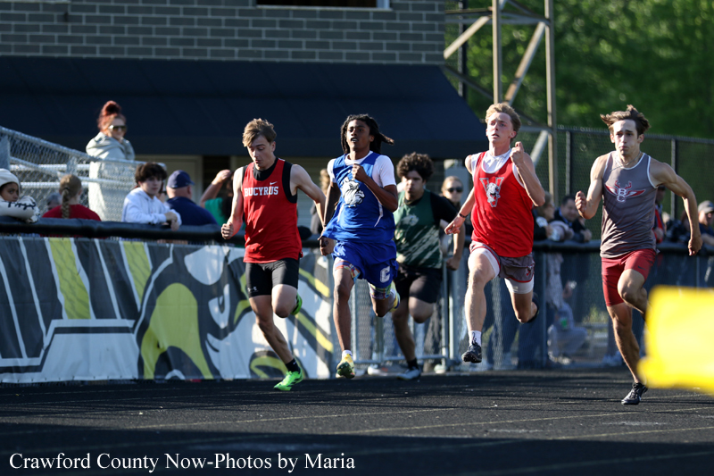 Five male sprinters race on a black track, wearing red, blue, and gray uniforms, with spectators behind the fence.