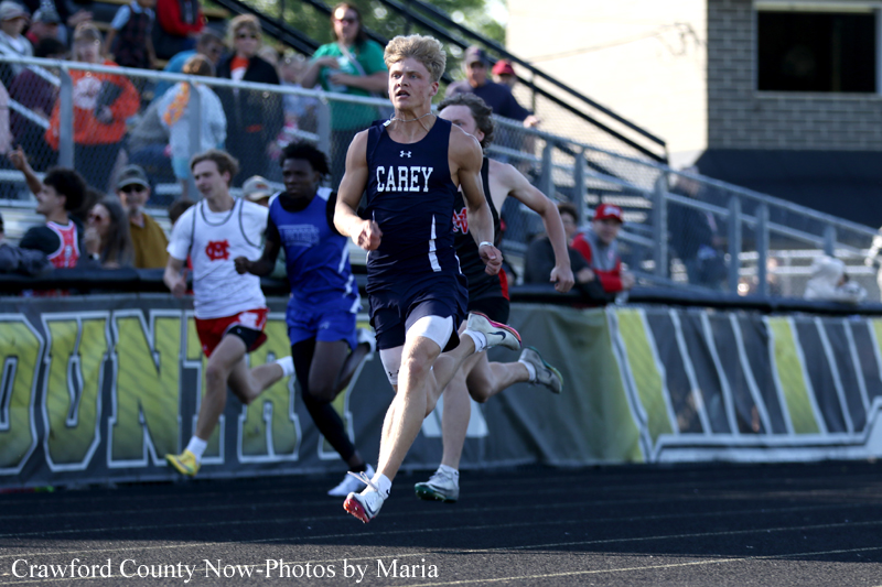 Male runner in a navy Carey uniform leads a sprint on an outdoor track, with other competitors and spectators in the background.