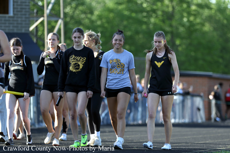 Group of young female athletes walking on a track, wearing black and gray shirts with eagle logos, outdoors at a meet.