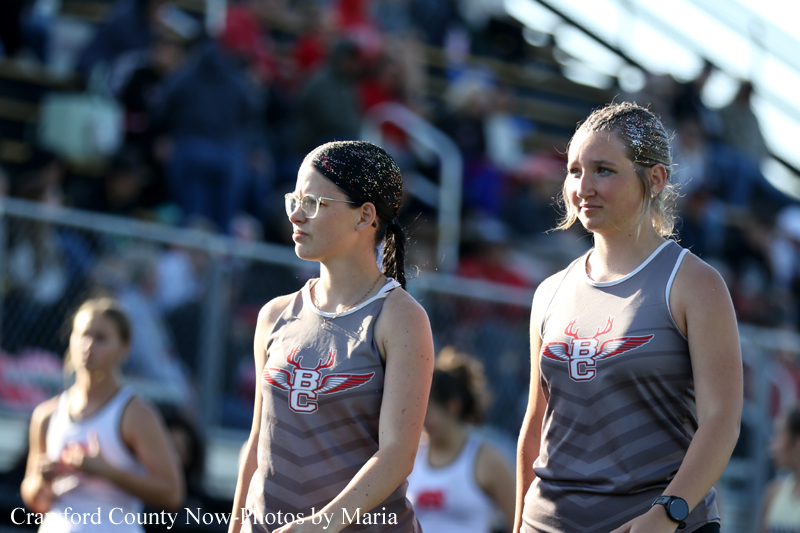 Two female runners in matching gray singlets with a red winged logo stand on a track, with a crowded stadium in the background.