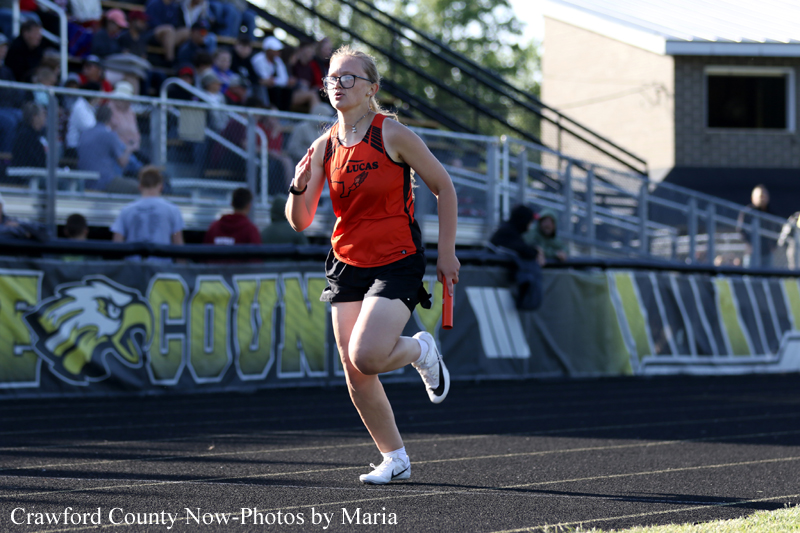 Female track sprinter in an orange jersey runs on a track, holding a red baton.