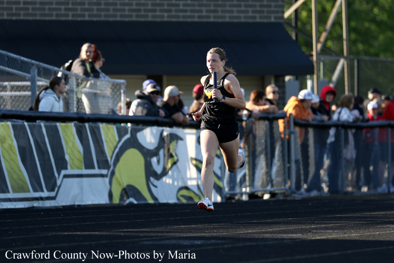 Female sprinter in a black uniform runs on an outdoor track as spectators line the fence behind her.
