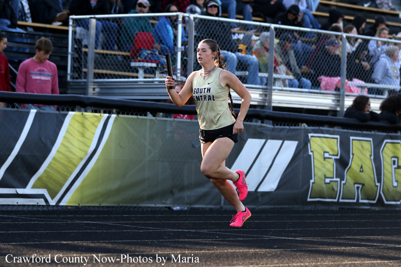 Female relay runner in a gold jersey labeled South Central, sprinting on the track with baton in hand, stands and spectators behind her.