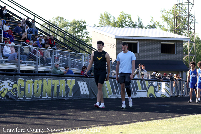 Two male athletes walk along a running track beside a crowded bleacher, with a county-themed banner in the background and a brick building behind them.