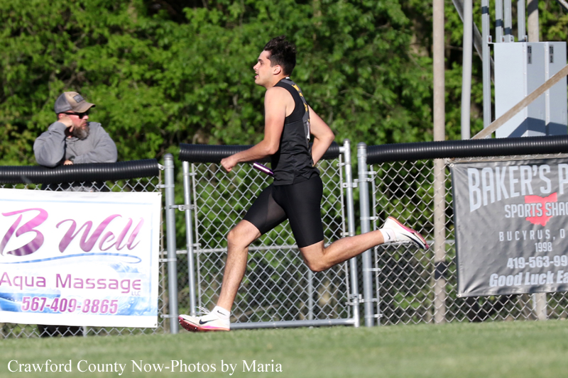 Male runner in black athletic gear sprints along a track beside a chain-link fence with banners in the background.