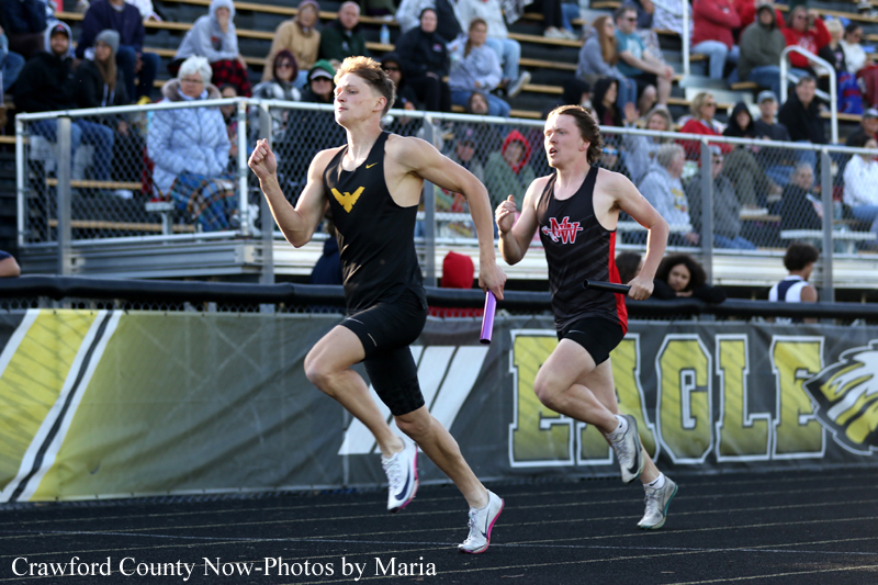 Two male relay runners sprint on a track, each holding a baton, with spectators in the stands behind.
