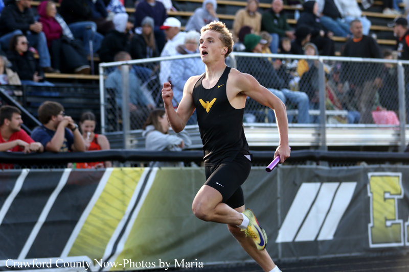 Male track athlete in a black uniform sprinting on a relay race, holding a purple baton, with spectators in the stands behind.
