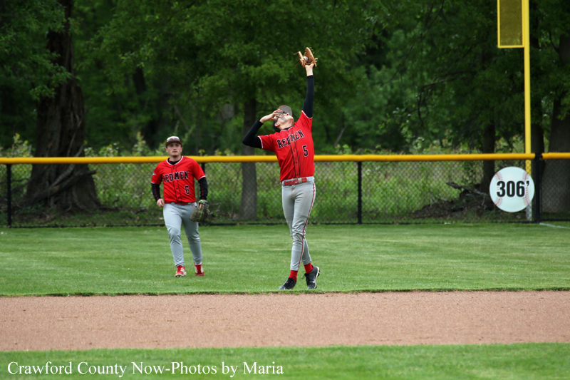 Two baseball players in red uniforms on a grassy field; one pitcher raises his glove to throw or catch, while a teammate watches, with a chain-link fence and yellow foul pole in the background and a 306' sign nearby.