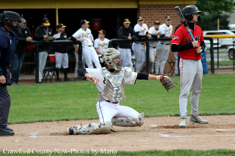 Youth baseball scene: catcher slides into home plate as batter in red watches; dugout and spectators in the background.